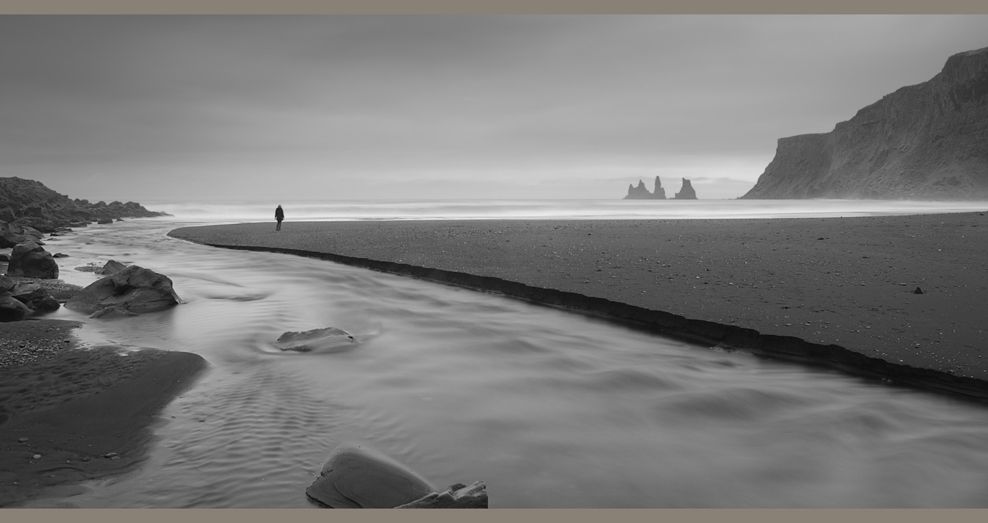 A figure stands on a wedge of black sand between river, sea and sky